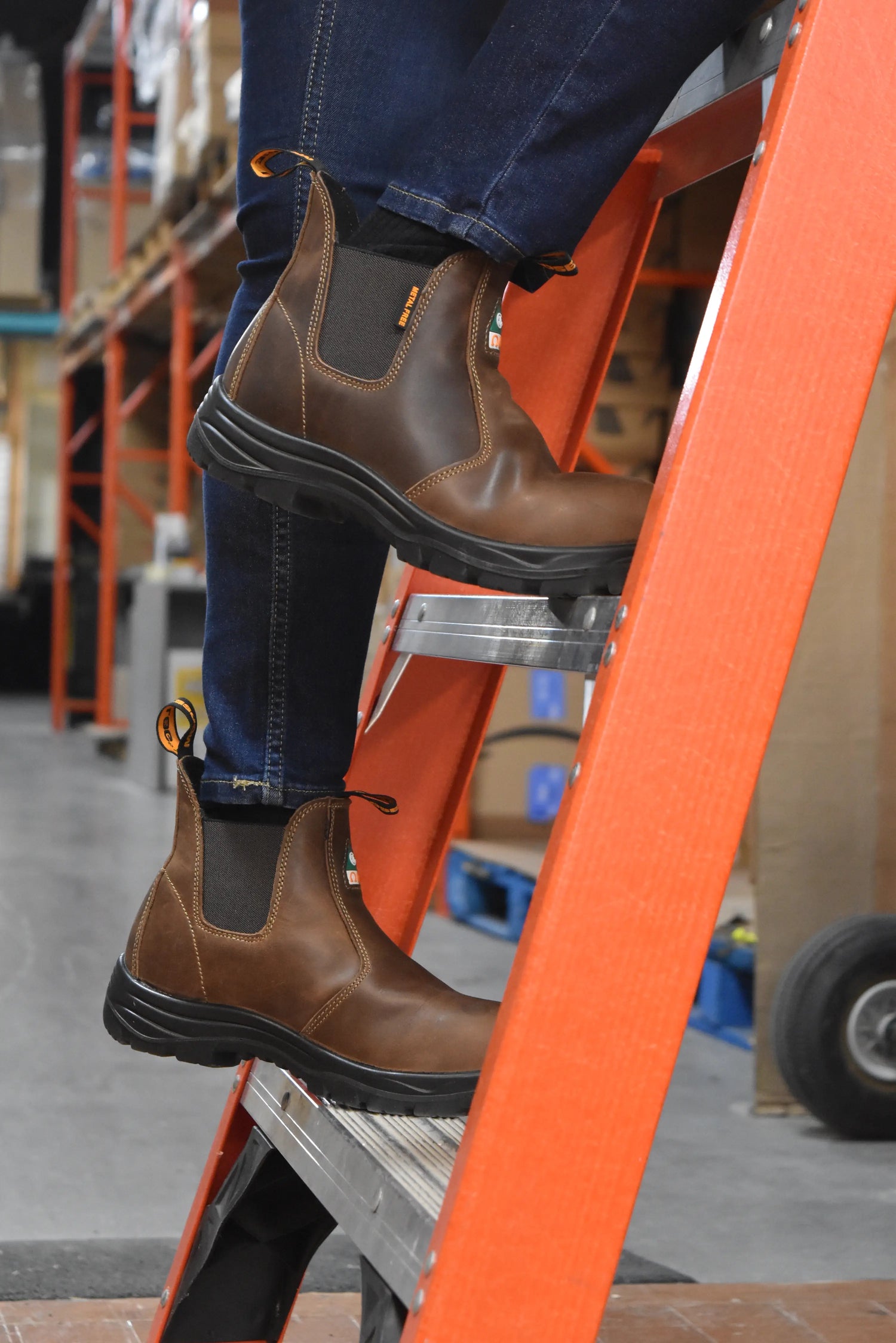 Woman wearing brown chelsea work boots on an orange ladder in a warehouse setting
