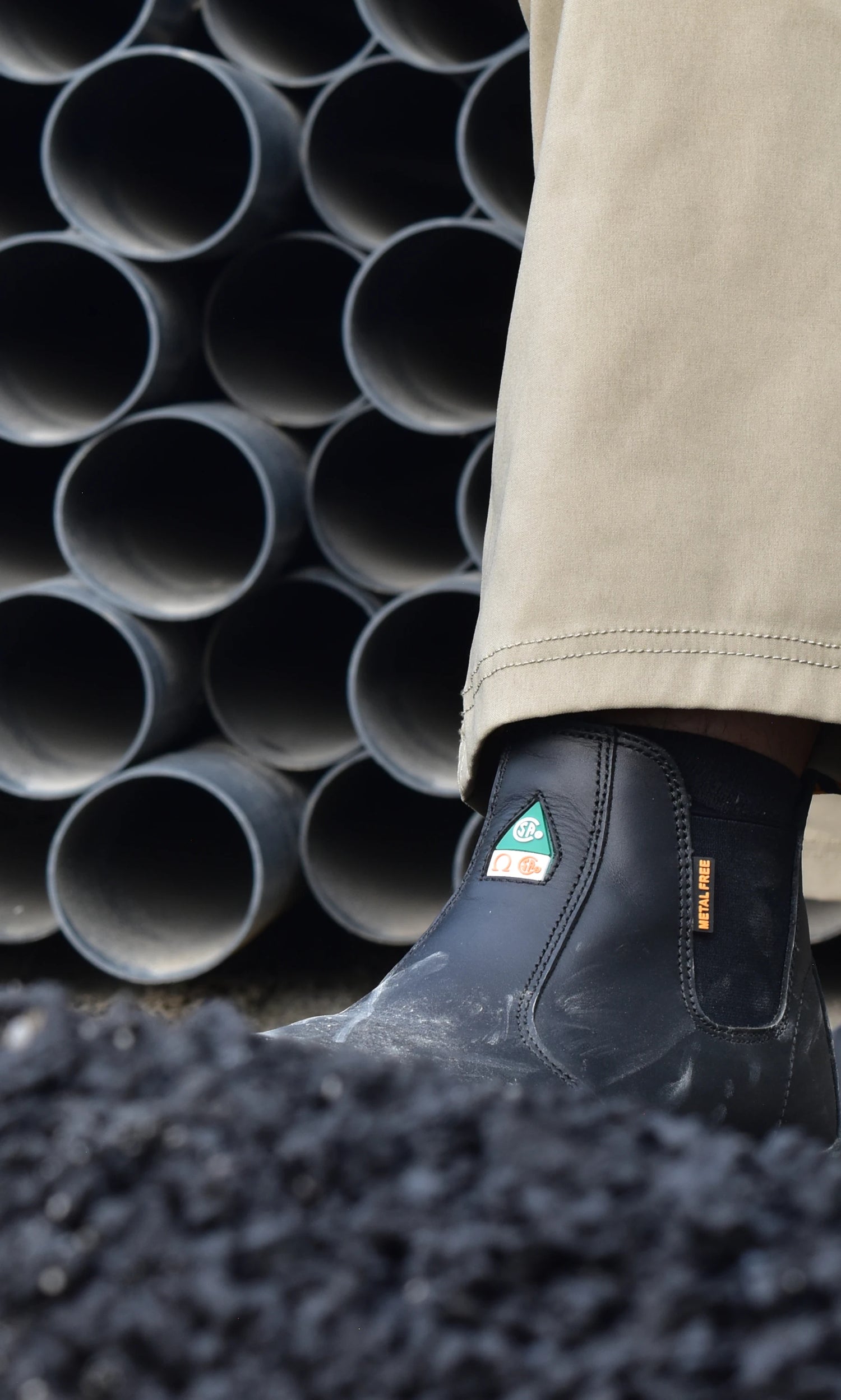 Person wearing black rubber boots with a CSA logo, standing on black asphalt with stacked pipes in the background.