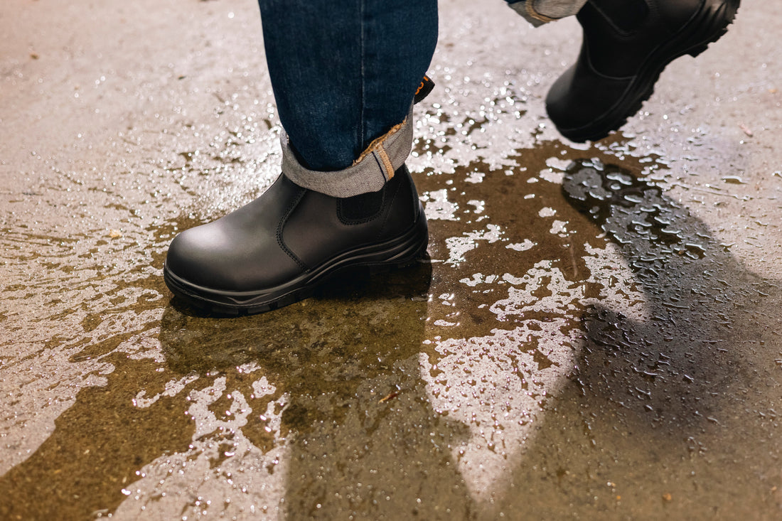 Black composite toe work boots on wet industrial floor, worn with rolled-up jeans
