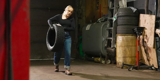 Woman in work boots lifting tire in industrial garage, surrounded by stacked tires and equipment