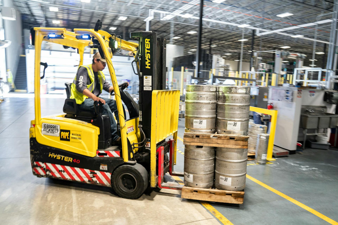 Worker driving yellow forklift moving metal kegs on pallet in industrial warehouse