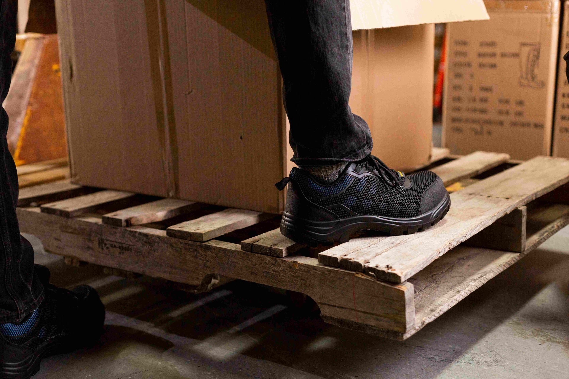 Person wearing black shoes on a wooden pallet with cardboard boxes in the background