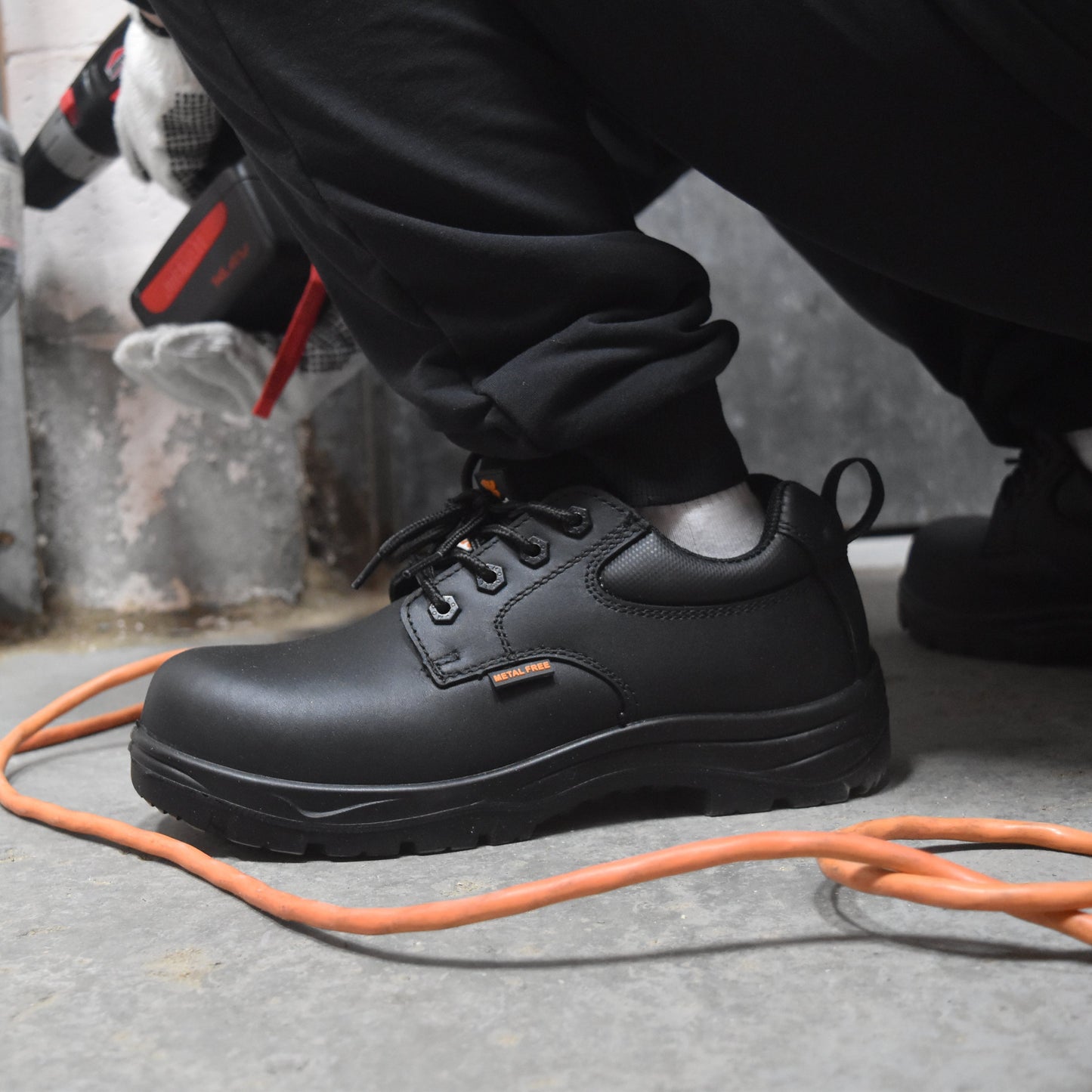 A person wearing a pair of black leather work boots with white gloves, kneeling on a concrete floor next to an orange extension cord.
