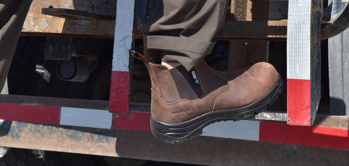 A person wearing brown slip-on work boots and dark pants steps onto the metal ladder of a truck, with the truck bed and Ontario license plate visible in the background.