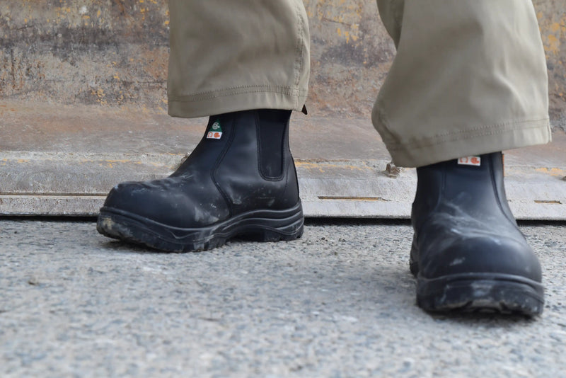 Black rubber boots worn with beige pants on a concrete surface.