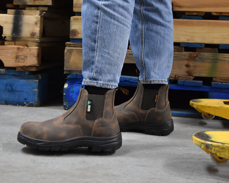 Person wearing brown work boots and blue jeans standing on a concrete floor with wooden pallets in the background.