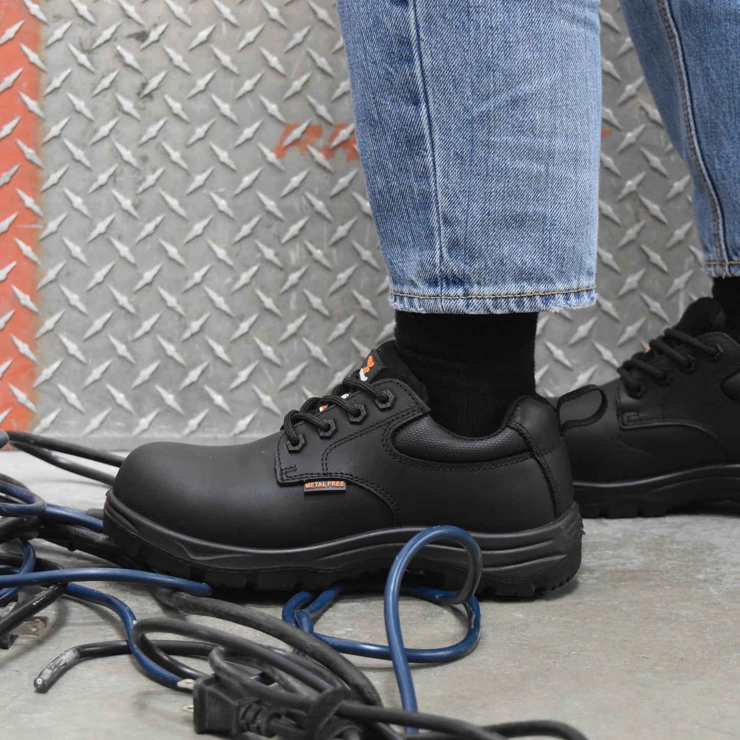 Black leather work boots worn with blue jeans and black socks, standing on a concrete floor in front of a diamond plate wall.