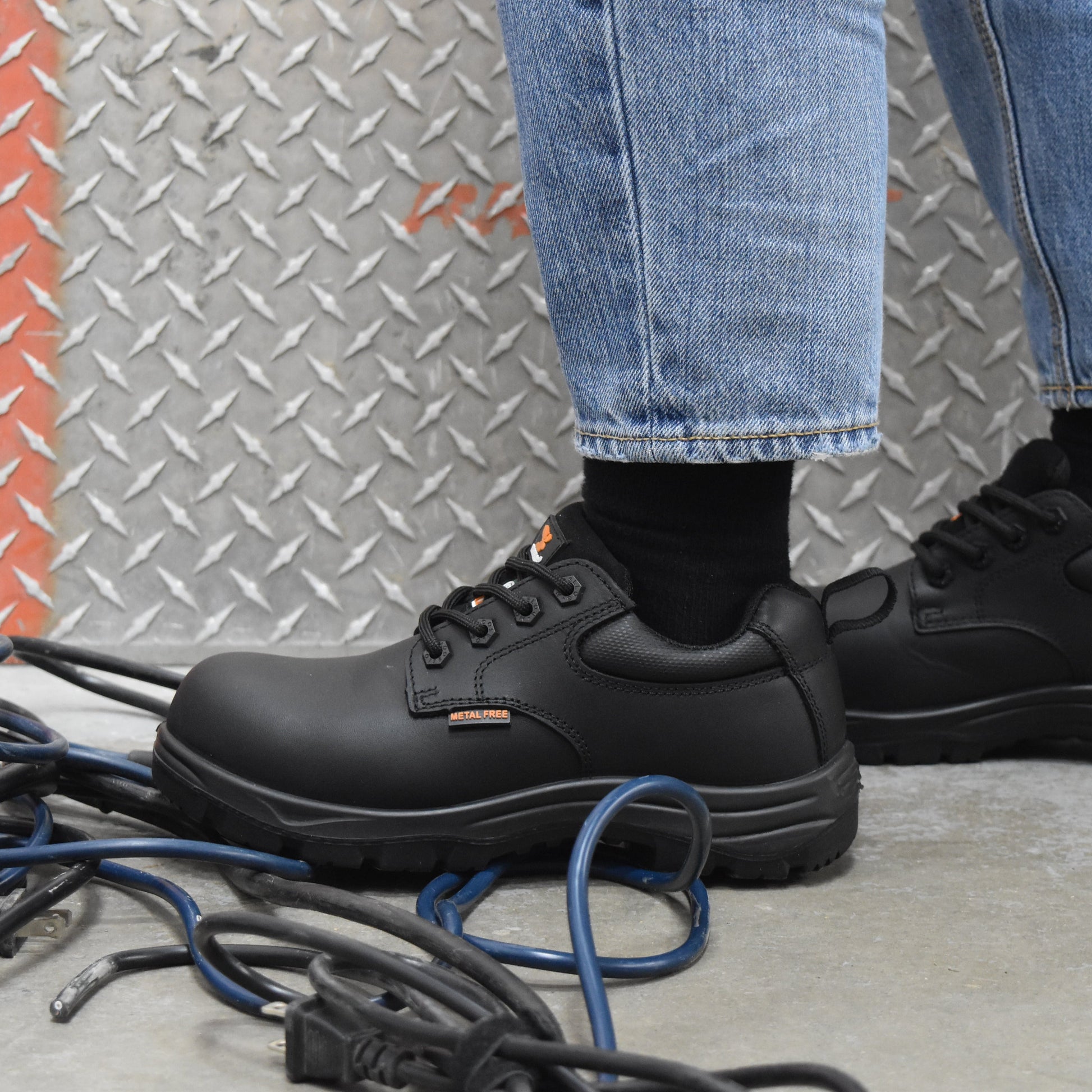 Black leather work boots worn with blue jeans and black socks, standing on a concrete floor in front of a diamond plate wall.