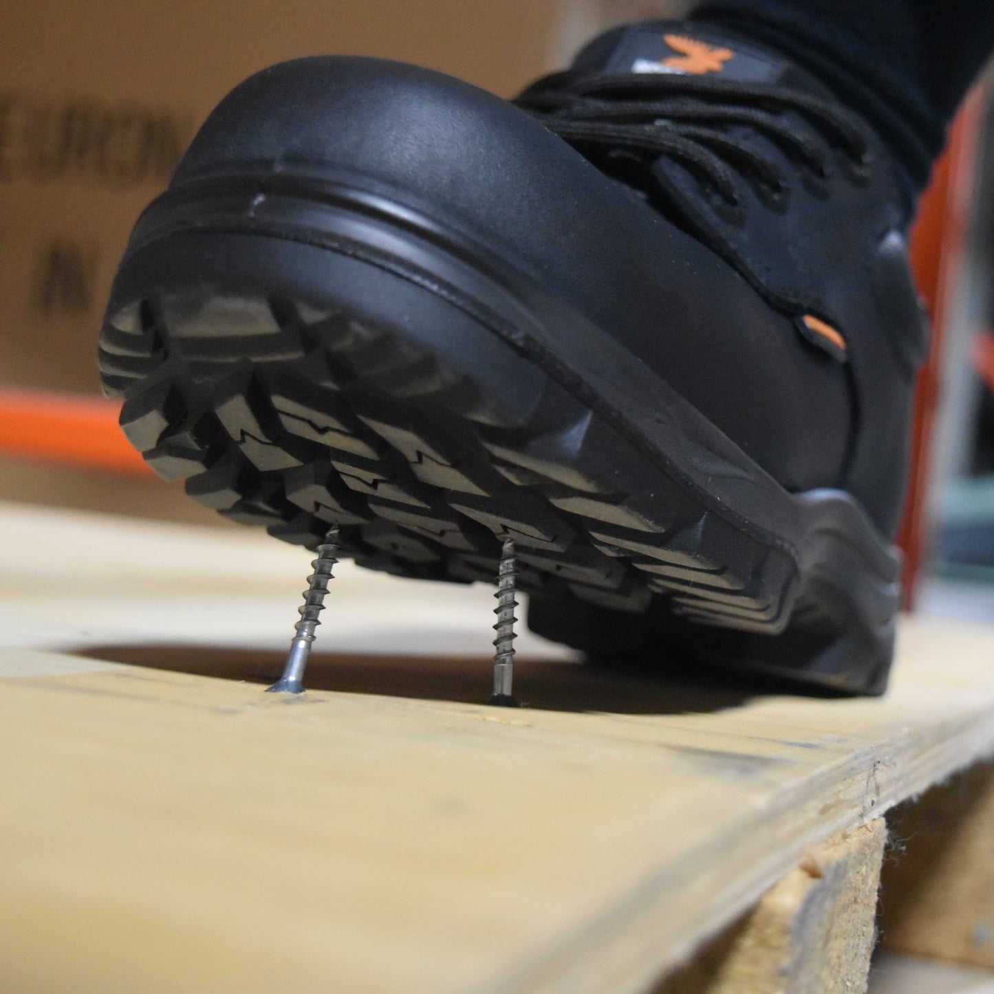 A close-up of a black safety boot stepping over two large metal screws placed on a wooden board, with a blurred indoor background.