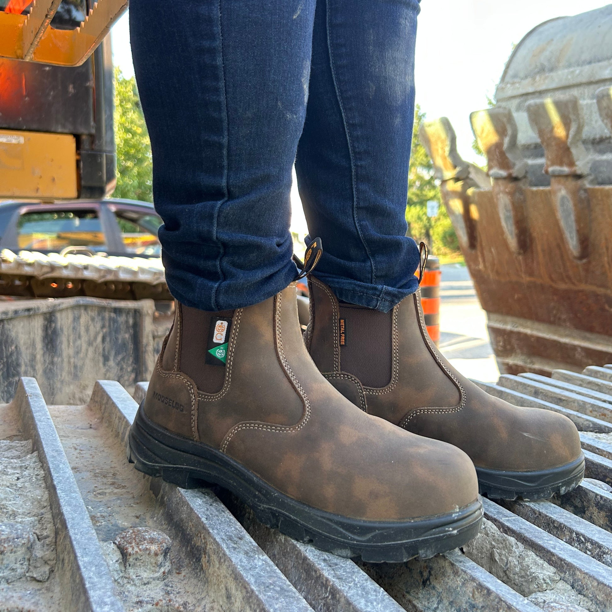 A person wearing dark brown leather safety work boots with elastic side gore panels and dark blue jeans, standing on a concrete grating at an outdoor construction site.