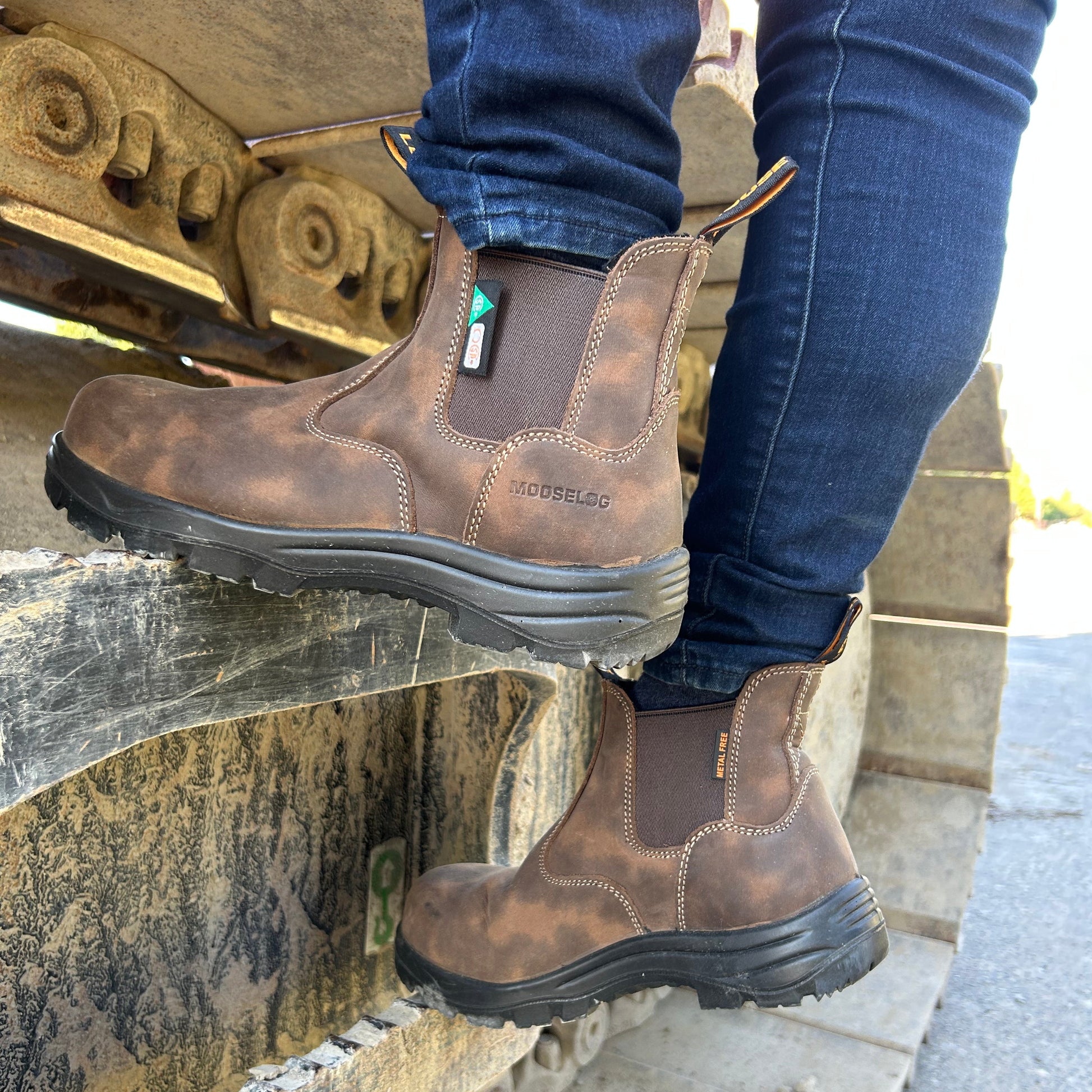 A person wearing brown leather work boots with dark elastic side panels and dark soles, standing on heavy machinery tracks outdoors.
