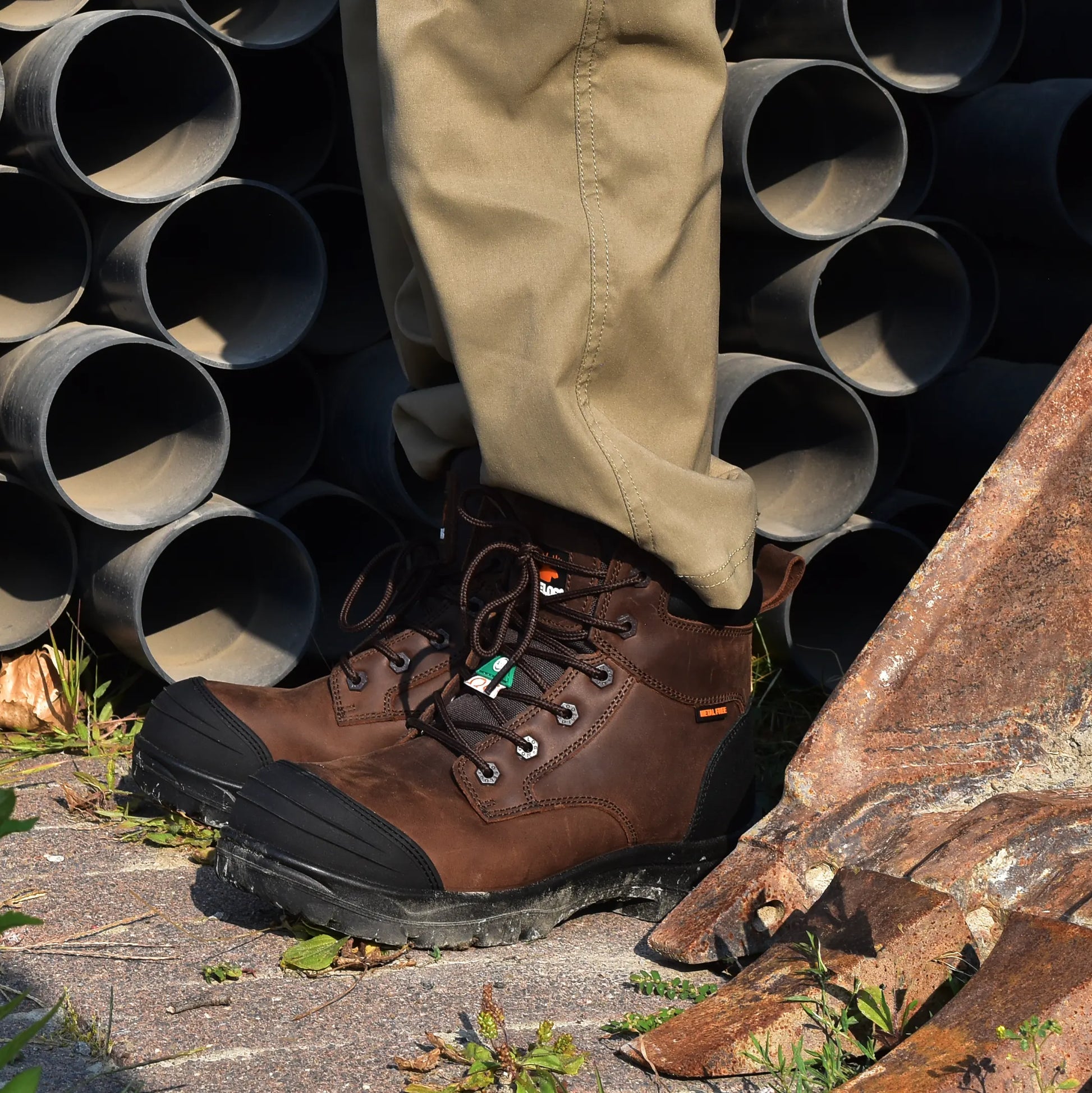 Brown work boots and beige pants with a background of stacked pipes and rocks.