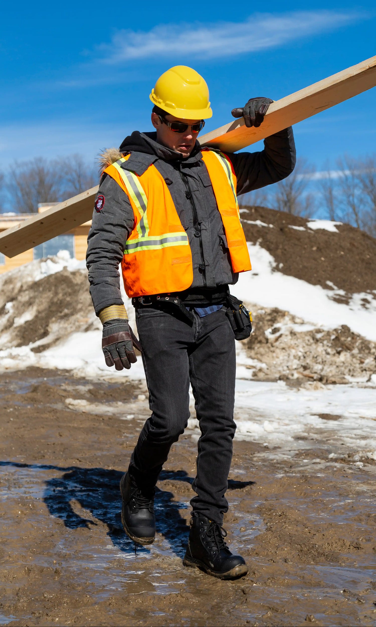 PPE man in safety boots carrying a wooden board on a construction site with snow and blue sky in the background.