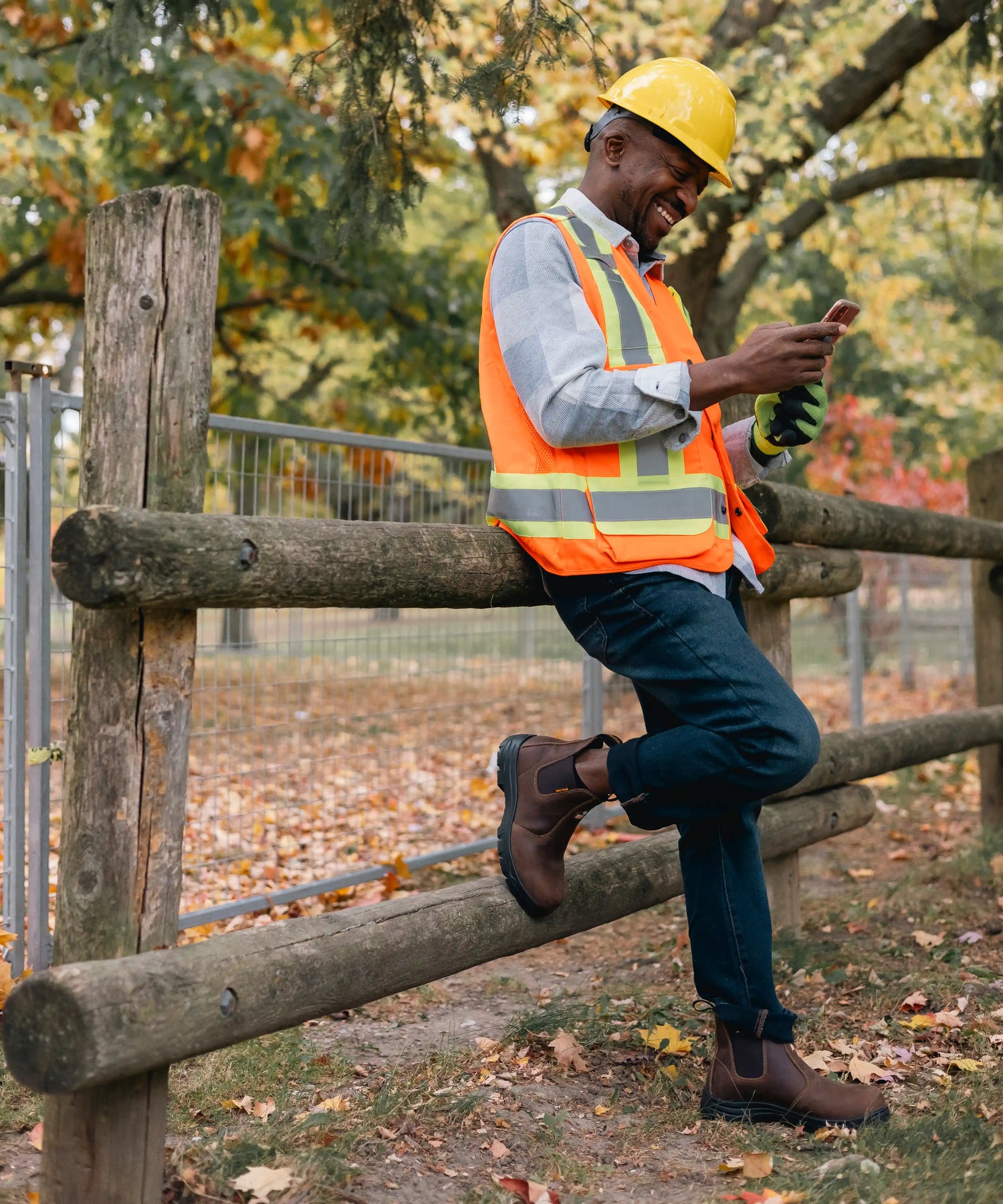 Person in safety vest, MooseLog boots, and hard hat sitting on a wooden fence outdoors